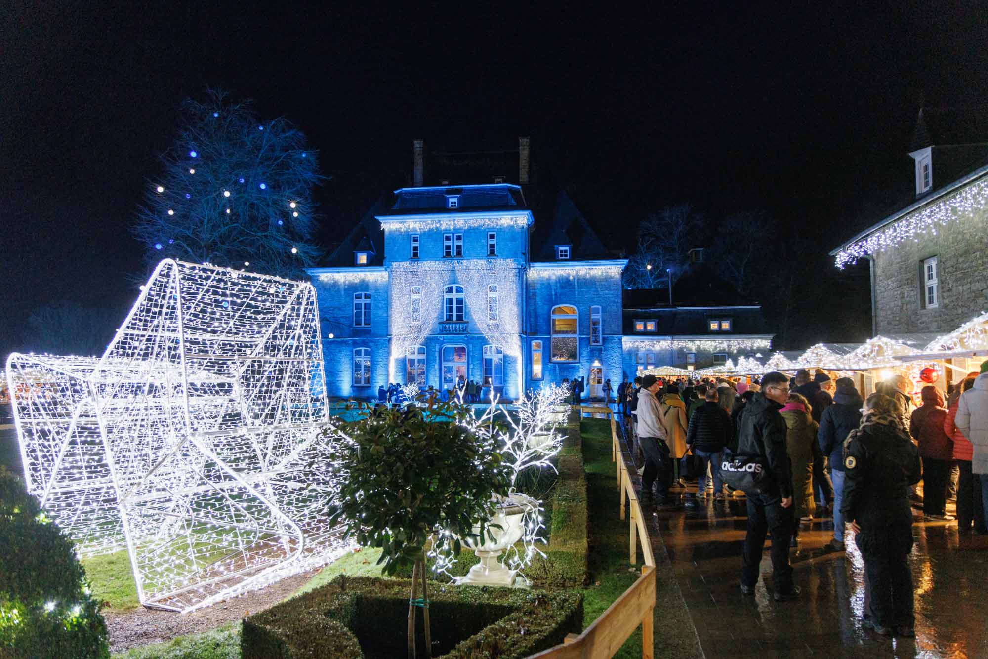 Marché de Noël de Ciney de nuit avec décorations lumineuses et visiteurs devant un bâtiment éclairé en bleu.