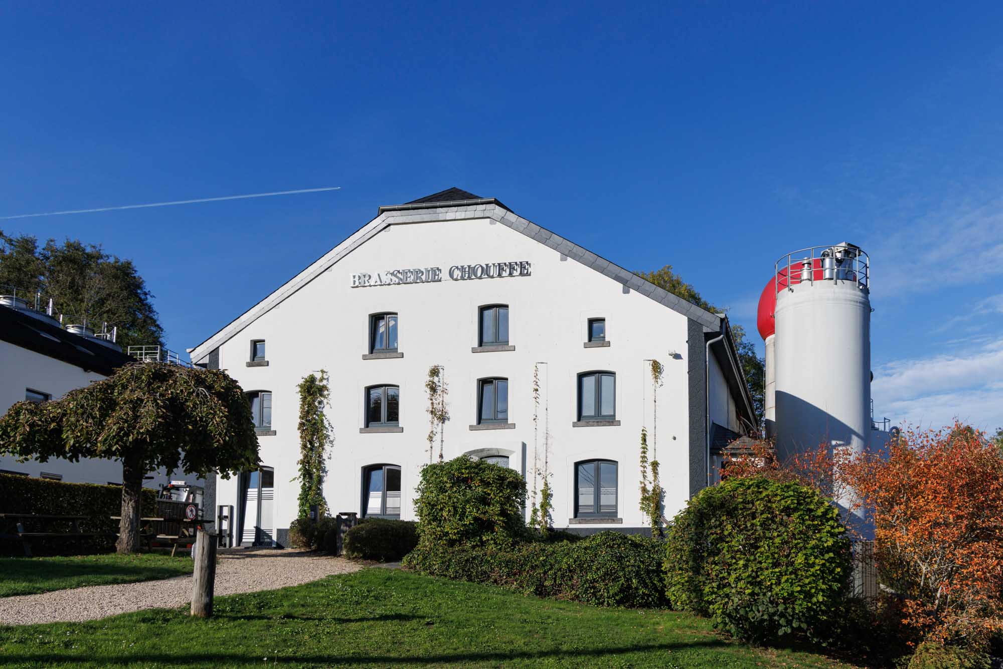 Gevel van de Brasserie Chouffe in Achouffe met het witte gebouw en de brouwerij-installaties onder een blauwe lucht.